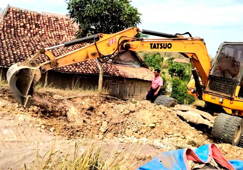 Warga Dusun Gunung, Desa Pasarenan, Kecamatan Kedungdung, Kabupaten Sampang berada di sekitar alat berat yang digunakan untuk memperbaiki jalan rusak dengan swadaya. (IBNU ANSORI / KLIKMADURA)